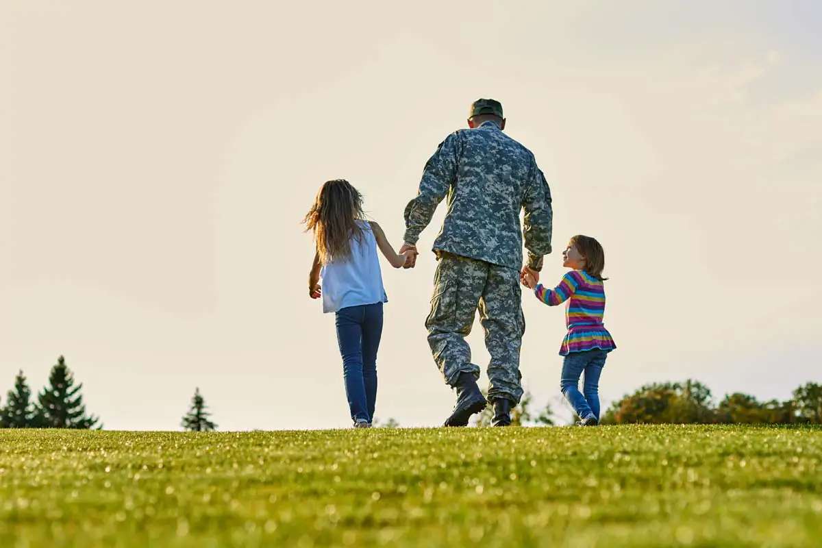 Service member walking hand-in-hand with two children across a grassy field