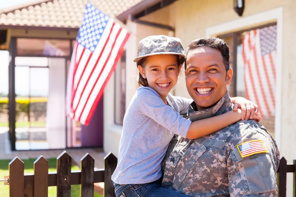 Service member holding a child in front of their home with an American flag